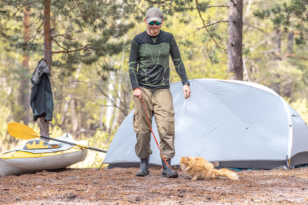 tourist on the river bank next to a kayak and a tent walks a ginger red cat on a leash